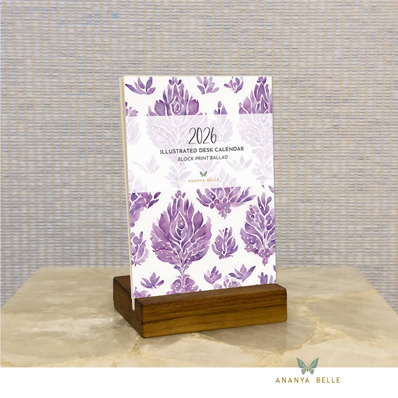 May include: A 2026 desk calendar with a purple floral design, displayed in a wooden stand. The calendar's cover features the text "2026 Illustrated Desk Calendar Block Print Ballad" and the brand name "Ananya Belle".