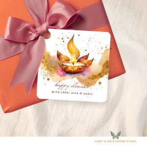 May include: A square gift tag with a watercolor illustration of a lit diya lamp, with the text "happy diwali" and "with love, Alia & Sahil". The tag is attached to a wrapped gift with a pink bow.