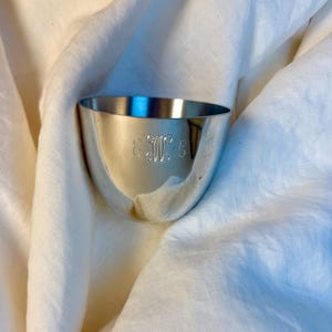 以下が含まれることがあります： 丸みを帯びた形の小さな銀色のボウル。ボウルには前面にイニシャルが刻まれています。背景は白のテクスチャード加工された生地です。