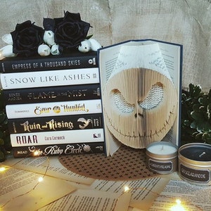May include: A stack of books with titles like "Empress of a Thousand Skies" and "Snow Like Ashes" next to a book with a carved owl design. Two small candles in metal tins are labeled "Currently Reading" and "GoogeAranddie". Black roses and white flowers are arranged on top of the books.