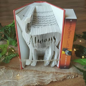 Peut inclure: Un livre plié en forme de cerf, posé sur une surface en toile de jute. Le livre est intitulé "Harry Potter et le Prince de Sang-Mêlé" par J.K. Rowling.