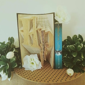 May include: A book folded into the shape of the Harry Potter symbol, the Deathly Hallows, with a copy of "Harry Potter and the Prisoner of Azkaban" by J.K. Rowling in front of it. The book is surrounded by white flowers and green plants.