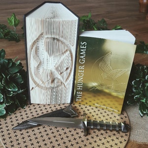 May include: A book titled "The Hunger Games" by Suzanne Collins, with a gold cover featuring a stylized bird design. The book is next to a book folded into a three-dimensional sculpture of a mockingjay symbol.