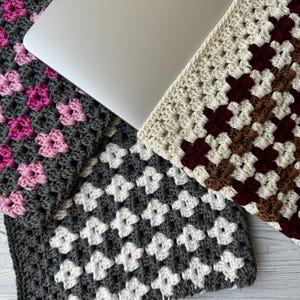 May include: A laptop rests on three crocheted laptop sleeves. The sleeves feature a granny square design in various color combinations: gray and pink, gray and white, and cream, brown, and burgundy. The handmade covers offer protection and style.