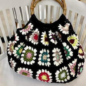 May include: A black crochet tote bag with a floral pattern in various colors, including white, red, yellow, green, and pink. The bag has two wooden handles. The bag is sitting on a white chair.