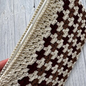 May include: Crocheted case in cream, brown, and burgundy. The case features a granny square pattern and a cream-colored border. The case is designed to hold a rectangular object, such as a tablet or notebook.