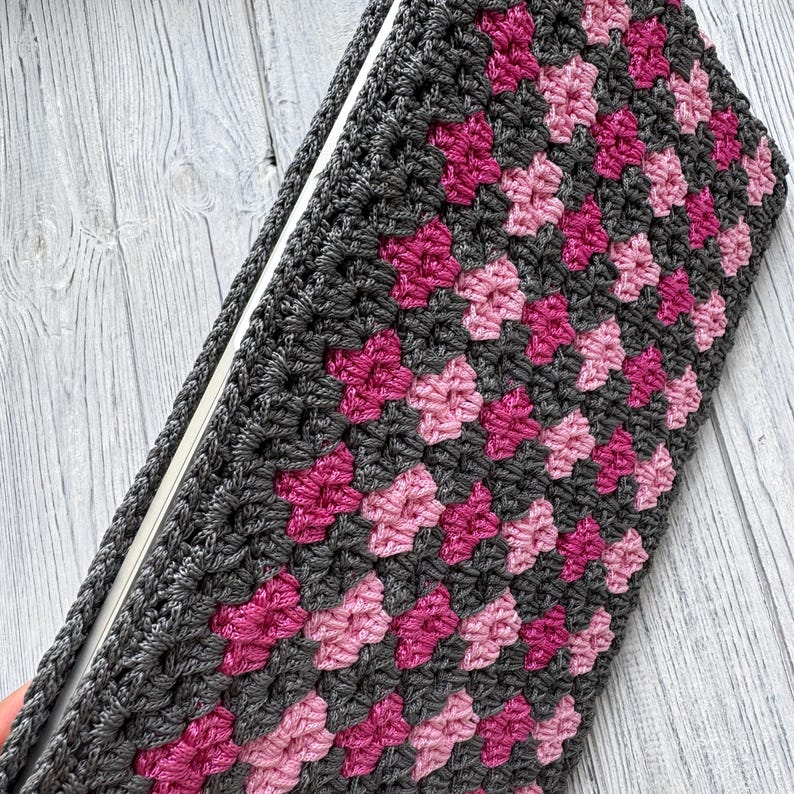 May include: A handmade crochet notebook cover in a granny square pattern. The cover features a gray base with pink and fuchsia floral motifs. The notebook is open, revealing white pages. A gray crocheted strap is visible.