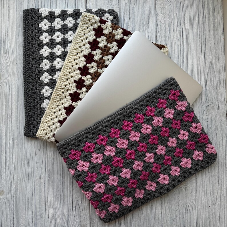 May include: A collection of crocheted laptop sleeves in various colors. The sleeves feature a granny square design in gray, white, cream, brown, and pink. A silver laptop is partially visible, resting inside one of the sleeves. The sleeves are arranged on a light wood-grain surface.