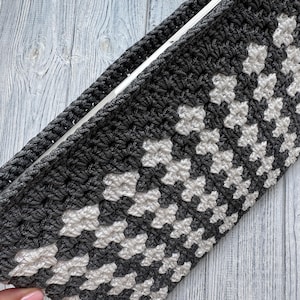 May include: A handmade crochet case in a geometric pattern of gray and off-white. The case is rectangular with a solid gray border and a textured, open weave design. The crochet work appears sturdy and well-crafted.