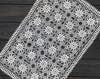 May include: White, rectangular crochet doily with a floral pattern. The doily features a detailed openwork design with repeating flower motifs and a scalloped border. The handmade textile is placed on a dark wood surface.