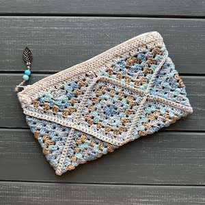May include: A rectangular crochet pouch featuring a granny square pattern in shades of light blue, beige, and cream. The top edge is finished with a cream border and a zipper closure adorned with a metal leaf charm and two small blue beads.