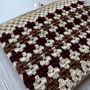 May include: A crocheted laptop sleeve in a granny square pattern. The sleeve features a repeating pattern of cream, brown, and burgundy squares. The sleeve is designed to fit a laptop and provides a protective layer.