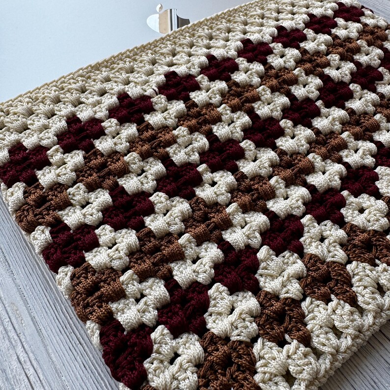 May include: A crocheted laptop sleeve in a granny square pattern. The sleeve features a repeating pattern of cream, brown, and burgundy squares. The sleeve is designed to fit a laptop and provides a protective layer.