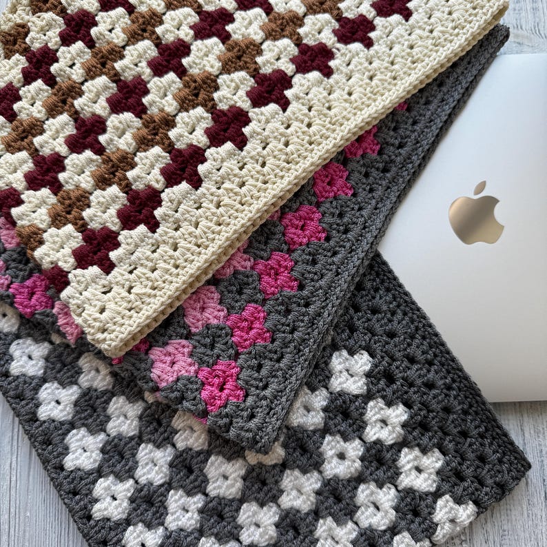 May include: Three crocheted laptop sleeves in a granny square pattern. The top sleeve features cream, brown, and burgundy squares. The middle sleeve is gray with pink flower squares, and the bottom sleeve is gray with white flower squares. A silver laptop is partially visible.
