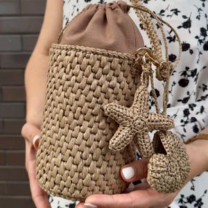 May include: A beige woven handbag with a drawstring closure and a matching shoulder strap. The bag features a starfish and seashell charm. The bag is cylindrical in shape and appears to be handmade.