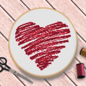 Pode incluir: Um padrão de ponto de cruz em forma de coração vermelho em um aro de bordado de madeira. O coração é feito de pequenos pontos vermelhos e brancos, criando um efeito texturizado.