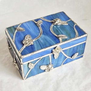 Peut inclure: Une boîte rectangulaire en verre teinté composée de panneaux de verre marbré bleu et blanc. L'extérieur est orné de détails en métal argenté soudé représentant des vignes sinueuses, des feuilles et de petits boutons de rose sur le couvercle.