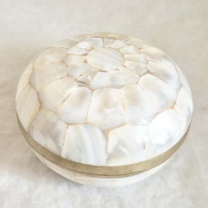 Peut inclure: Une boîte ronde décorative avec un couvercle en mosaïque de nacre. La boîte est entourée d'une bande de couleur laiton. La mosaïque est composée de pièces irrégulières, blanches et iridescentes.