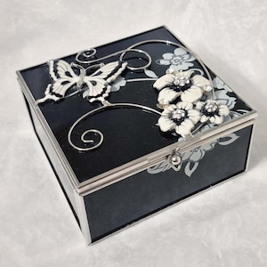 May include: Black jewellery box with a silver trim. The box is decorated with a white and silver butterfly and white flowers with crystal accents. The box has a hinged lid and a silver clasp.