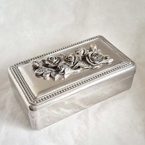 Puede incluir: Una caja rectangular plateada con tapa abisagrada. La tapa está adornada con un relieve floral detallado de rosas y hojas. La caja tiene un borde de cuentas y un acabado pulido, ideal para joyas.