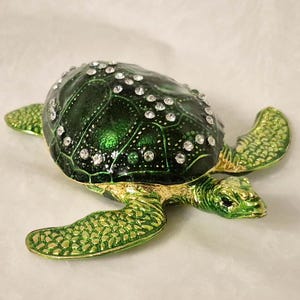 Può includere: Una statuetta decorativa a forma di tartaruga con un guscio smaltato verde scuro decorato con strass trasparenti scintillanti. La tartaruga presenta un corpo in metallo testurizzato color oro e verde con pinne e testa dettagliate.