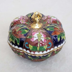 May include: A small, pumpkin-shaped trinket box with a floral design. The box is made of metal and has a gold-colored lid with a knob. The box is decorated with blue, green, pink, and brown enamel.
