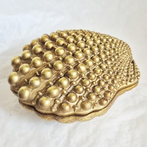 May include: A gold-colored decorative box shaped like a seashell. The exterior surface is covered in a dense pattern of raised, spherical beads of varying sizes, creating a textured, metallic finish that mimics the natural ridges of a shell.