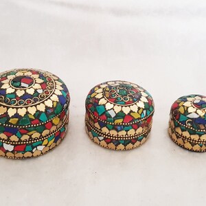 May include: Three round decorative boxes with intricate mosaic designs. The boxes are made of wood and are decorated with colorful glass pieces and gold accents. The boxes are different sizes, with the largest box in the back and the smallest box in the front.