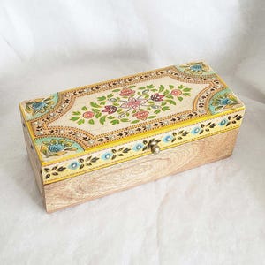 May include: Wooden rectangular box with a floral design on the lid. The box has a light wood base and a cream-coloured lid with a yellow border. The lid is decorated with pink, green, and blue floral patterns.