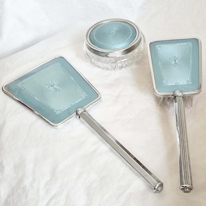 Puede incluir: Un juego de tocador vintage que incluye un espejo de mano, un cepillo y un tarro pequeño. El juego tiene marcos y mangos de metal plateado, con una incrustación de esmalte azul claro. El esmalte tiene un patrón de remolino decorativo.