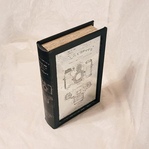 May include: A black book-shaped box with a silver metal front panel featuring a vintage camera patent drawing. The text on the front panel reads "L.R. camera".