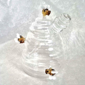 May include: A clear glass bong shaped like a beehive, featuring a spiralled mouthpiece. The bong has a lid and is adorned with three small bee figurines in brown and white. Set against a white, textured backdrop.