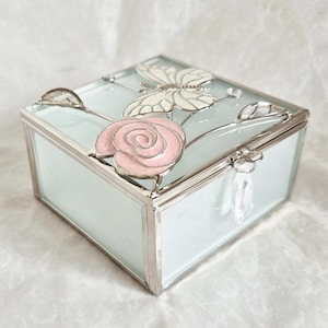 May include: A rectangular glass jewellery box with a light blue hue and silver trim. The lid features a pink rose and a butterfly design. The box has a small clasp with a clear crystal tassel.