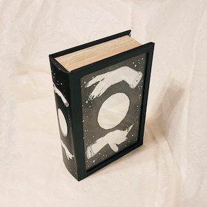 May include: Black box with a book-like design featuring a white illustration of hands reaching towards a moon. The box has a textured surface and a slightly raised lid.