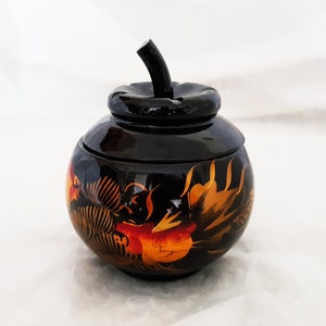 Könnte beinhalten: Eine schwarze Holzdose mit einem Deckel in Apfelform. Die Dose ist mit handbemalten goldenen und roten Blumenmustern verziert.