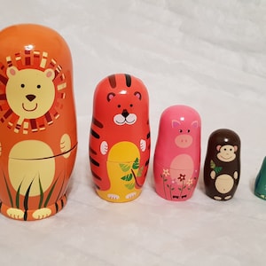 May include: A set of five wooden nesting dolls featuring animal designs. The largest is an orange lion, followed by a red tiger, a pink pig with floral details, a brown monkey, and a small green bird. Each doll has a smooth, painted finish.