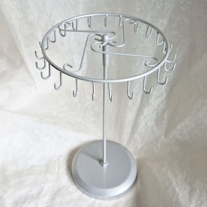 Puede incluir: Un expositor de joyas de metal plateado. El expositor tiene una parte superior circular con múltiples ganchos para colgar collares o pulseras. Cuenta con un poste de soporte central y una base redonda para mayor estabilidad. Diseñado para exhibir joyas.