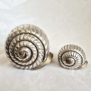 Op de afbeelding: Twee zilverkleurige, schelpvormige decoratieve objecten. De grotere schelp heeft een spiraalvormig ontwerp met een messingkleurig accent. De kleinere schelp weerspiegelt het ontwerp. Beide schelpen staan op een wit oppervlak.
