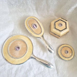 Könnte beinhalten: Ein Set aus vier Vintage-Schminkutensilien, darunter zwei Handspiegel, ein kleines Kompakt und eine sechseckige Box. Die Spiegel und die Box sind mit einem hellgelben Hintergrund, einem zarten Muster und einem einzigen lila Strassstein in der Mitte verziert.