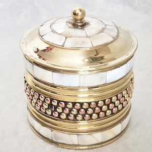 以下が含まれることがあります： 真珠貝のタイルが埋め込まれた蓋が付いた丸い真鍮製の容器。容器の中央には金と銅のビーズのバンドがあります。