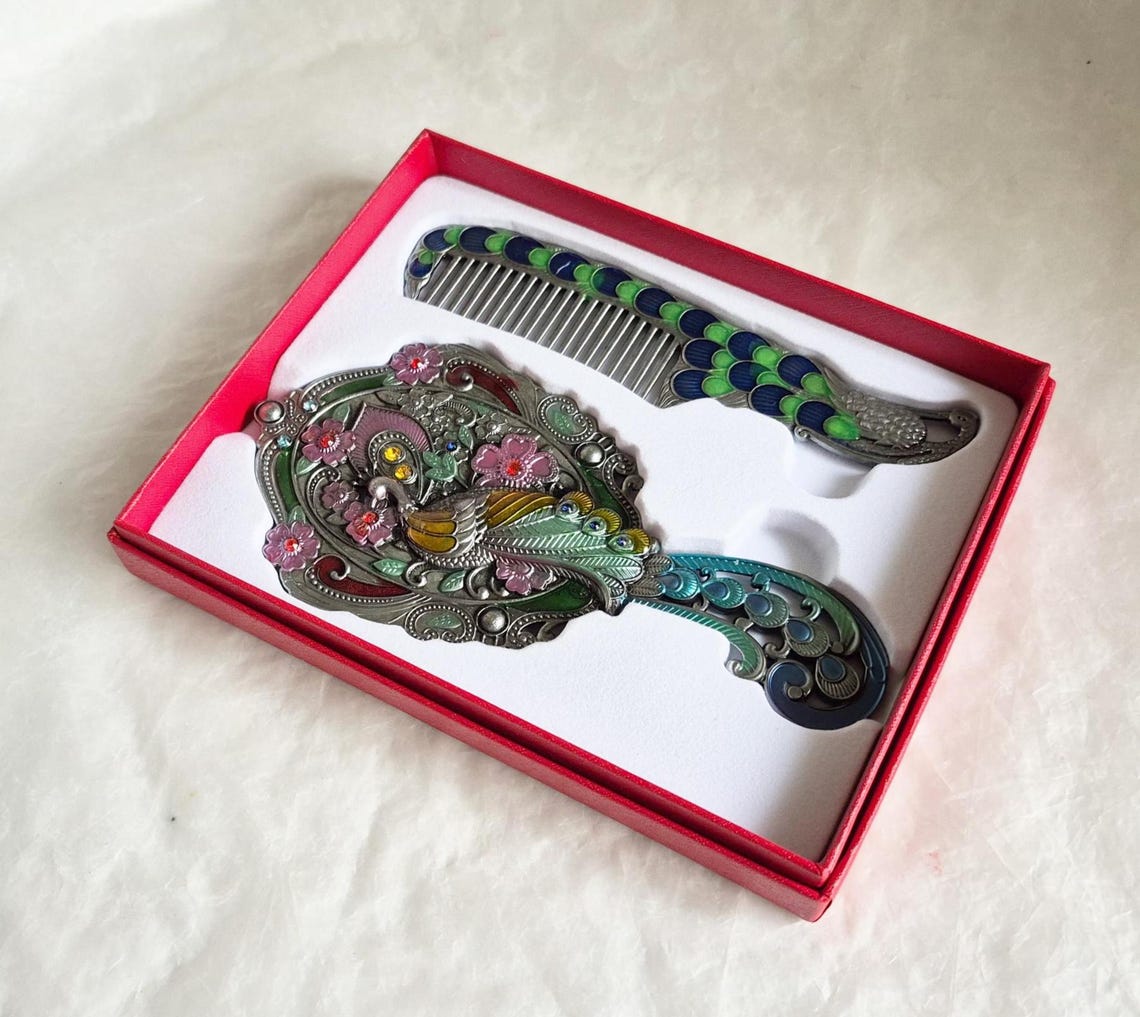Top view of mirror and comb from boxed vintage style enamelled hand mirror and comb, intricate teal, pink and silver peacock diamanté patterned fascia.