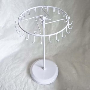 Op de afbeelding: Een witte metalen sieradenstandaard. De standaard heeft een cirkelvormige bovenkant met kleine haken om kettingen of armbanden op te hangen. Het heeft een centrale verticale paal en een ronde basis voor stabiliteit. De standaard is ontworpen om sieraden te presenteren.