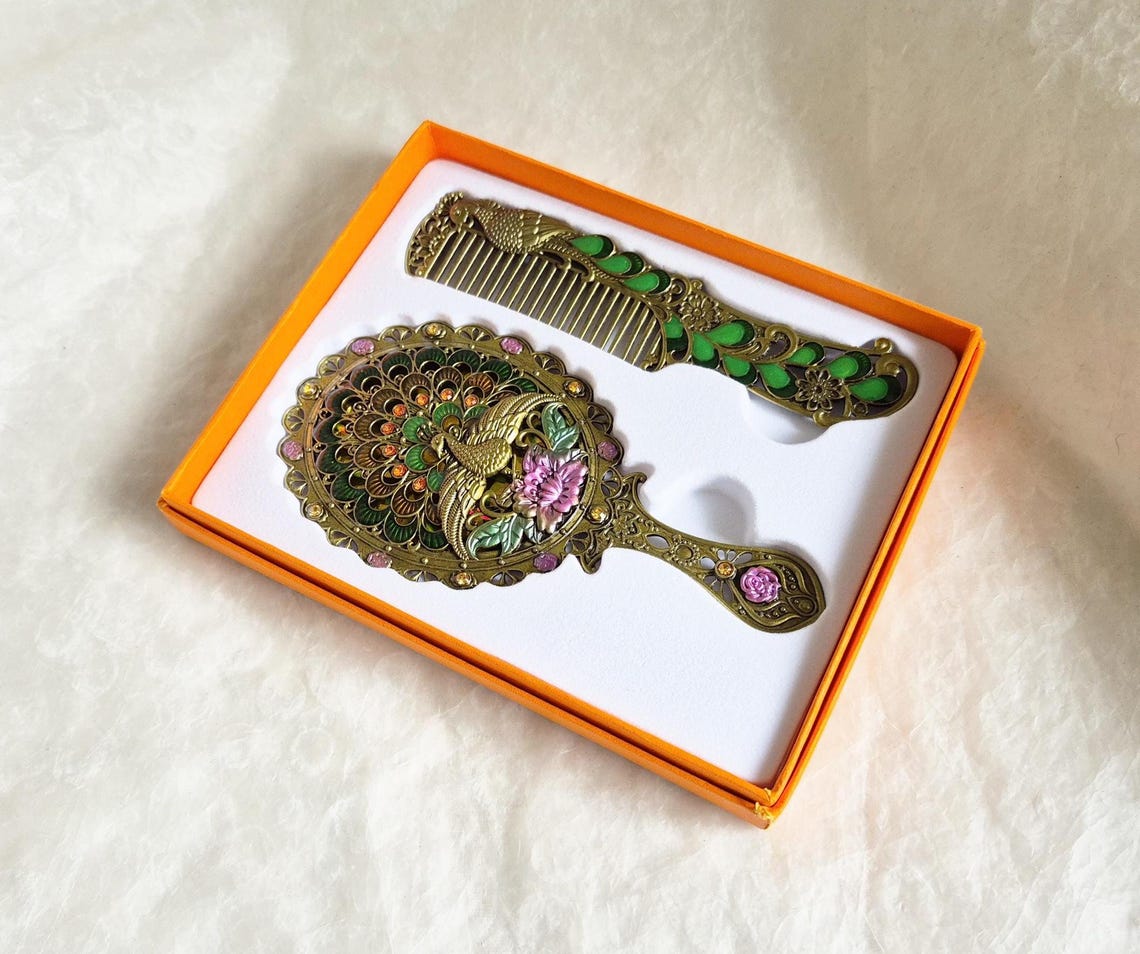 Top view of items inside box from vintage style enamelled hand mirror and comb, intricate green, pink and brass Peacock diamanté patterned fascia.
