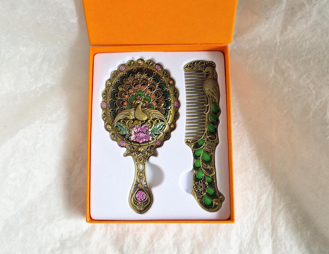 Top view of items inside box from vintage style enamelled hand mirror and comb, intricate green, pink and brass Peacock diamanté patterned fascia.
