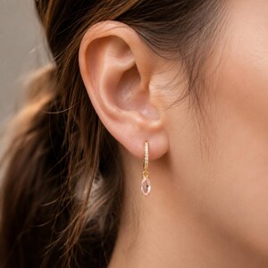 May include: Gold hoop earrings with small clear stones and a dangling oval-shaped pink gemstone. The earrings are shown on a person's ear, with brown hair in the background. The earrings are a delicate and elegant jewelry item.