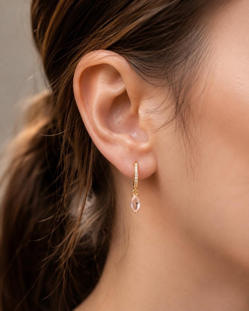 May include: Gold hoop earrings with small clear stones and a dangling oval-shaped pink gemstone. The earrings are shown on a person's ear, with brown hair in the background. The earrings are a delicate and elegant jewelry item.