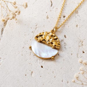 Può includere: Una collana color oro con un ciondolo circolare composto da due metà. La metà superiore ha una finitura dorata martellata e testurizzata, mentre la metà inferiore è un inserto liscio in madreperla bianca iridescente su una catenina sottile.