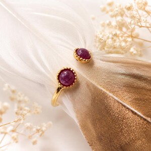 Peut inclure: Une bague ouverte en or ornée de deux pierres cabochons rondes d'un magenta profond, serties dans des chatons festonnés. L'anneau est lisse et poli, avec les deux pierres positionnées aux extrémités ouvertes, créant un design symétrique.