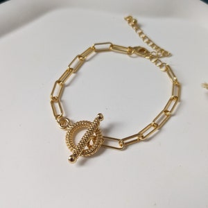 Peut inclure: Un bracelet en chaîne dorée avec un fermoir à bascule. Le fermoir est un cercle torsadé avec une barre qui le traverse.
