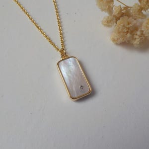 Peut inclure: Un collier en chaîne dorée avec un pendentif rectangulaire orné d'une incrustation de nacre blanche et d'un petit strass transparent.